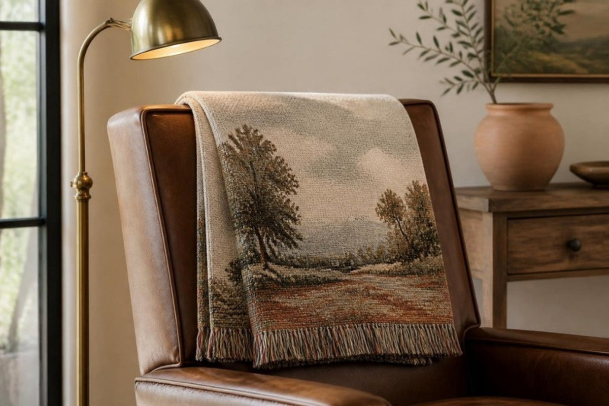 A woven cotton tapestry throw with a countryside landscape scene draped over the back of a leather armchair, with a brass floor lamp beside it.