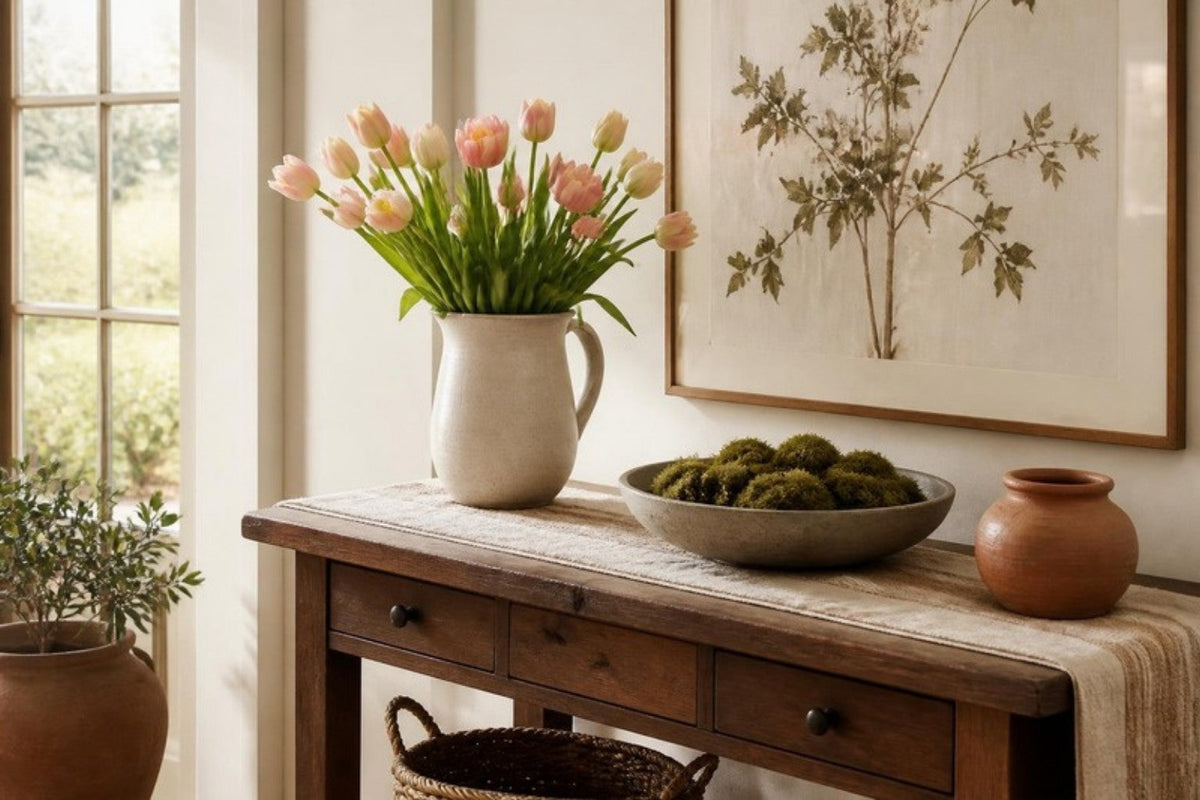 A wooden entry console with pink tulips in a white stoneware pitcher, a bowl of moss on a linen runner and a framed botanical print on the wall beside a sunlit window.
