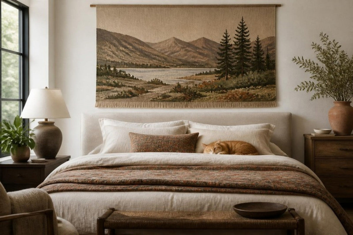 A large woven landscape tapestry of mountains and a lake hangs above a linen headboard while a ginger cat sleeps on the pillows and a folded quilt lies at the foot of the bed.