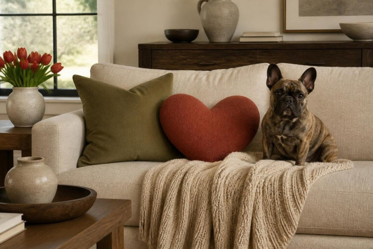 A French bulldog sits on a cream linen sofa beside a rust-red heart-shaped pillow and a knit throw, with a small vase of red tulips on the side table.