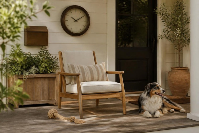 An Australian shepherd lies on porch boards beside a teak lounge chair with a striped cushion, with a wall clock, wall mailbox and a planter of greenery behind.